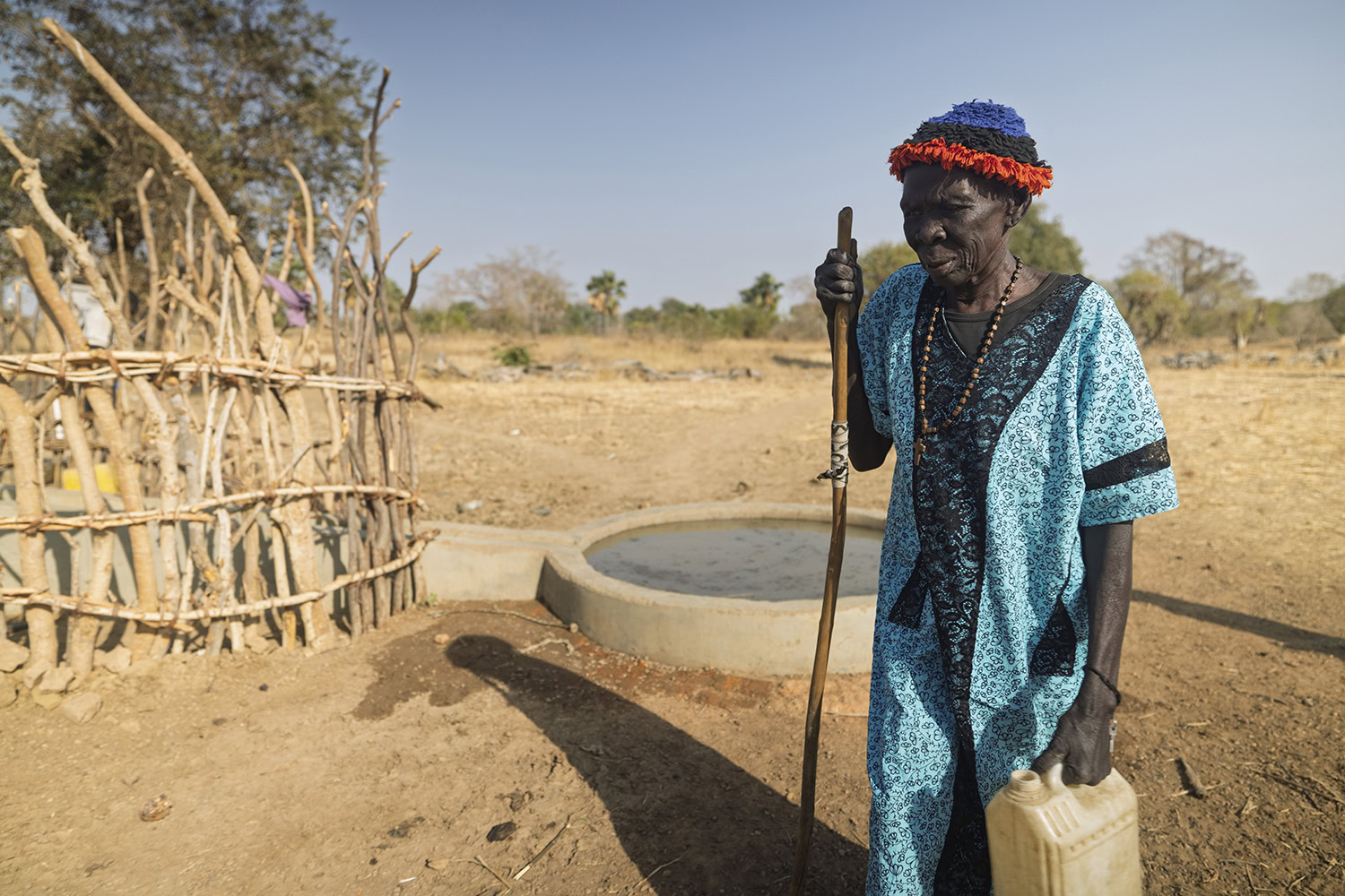 Een oudere vrouw in traditionele kleding haalt water uit een waterput in een dorpslandschap, onder een heldere blauwe hemel, met een houten hek op de achtergrond.