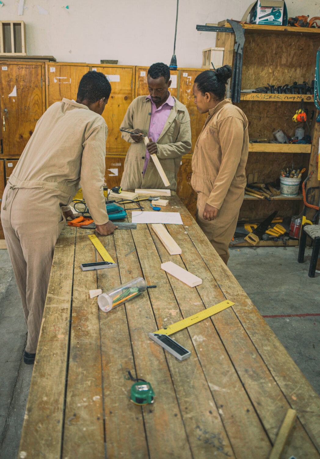 Drie mensen bespreken en ontwerpen nieuwe ideeën in een werkplaats, omringd door gereedschap en materialen voor het ontwikkelen van automatische concepten.