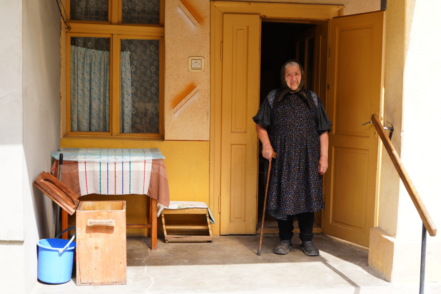Een oudere vrouw staat bij de ingang van haar huis, met een wandelstok in haar hand, in een warme en uitnodigende omgeving.