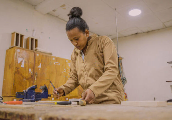 Een vrouw die geconcentreerd werkt aan een houten project in een werkplaats, met gereedschap en houtmaterialen op de tafel, onder goede verlichting.