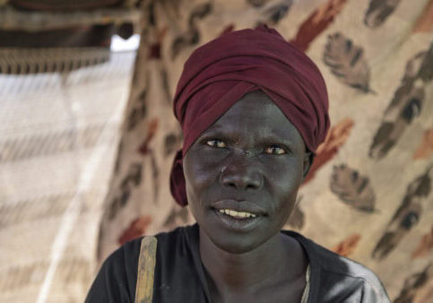 Een close-up van een vrouw met donkere huid en een rode hoofddoek, lachend en kijkend naar de camera, met een achtergrond van een tent en natuurlijke omgeving.