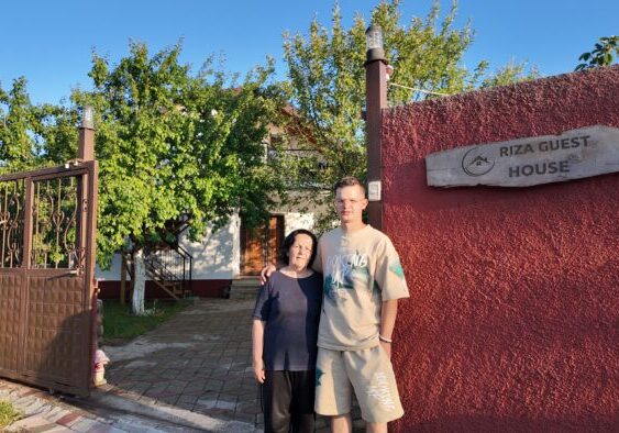 Een foto van een vrouw en een jonge man voor de ingang van Riza Guest House, omgeven door groene bomen en een heldere blauwe lucht.
